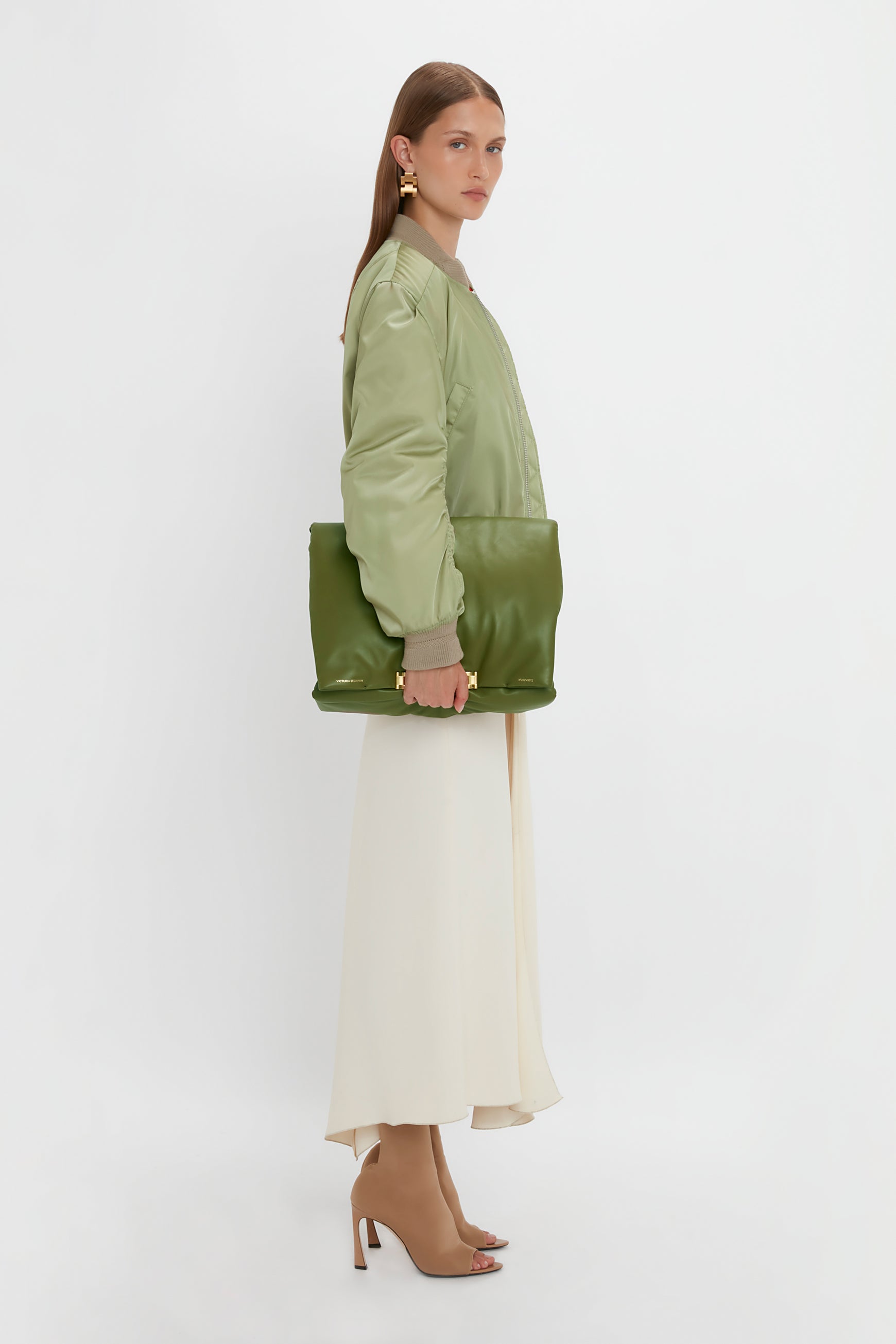 Woman in a green bomber jacket and long white skirt, holding a Victoria Beckham Puffy Jumbo Chain Pouch In Khaki Leather with an adjustable shoulder strap. She is wearing tan open-toe heels and gold earrings, standing against a plain white background.