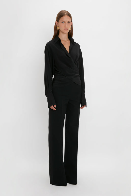 A woman stands against a plain white background wearing a black wrap-style blouse and Victoria Beckham Satin Panel Straight Leg Trouser. She has long, straight hair styled in a satin tuxedo braid and wears gold earrings.