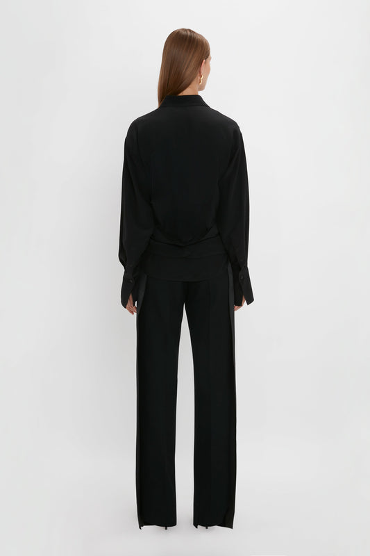 Woman in elegant Victoria Beckham black silk wrap front blouse and pantsuit viewed from the back, standing against a plain white background.