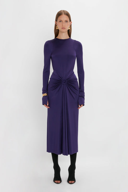 Person standing in the Victoria Beckham Long Sleeve Gathered Midi Dress In Ultraviolet, wearing black high-heeled shoes and gold earrings, against a plain white background.