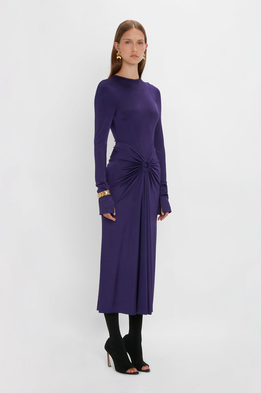A woman with long hair stands against a plain background, wearing a Victoria Beckham Long Sleeve Gathered Midi Dress In Ultraviolet with a circular gathered detail at the waist and black open-toe heels. She has gold earrings and a gold bracelet.