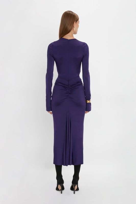 A woman stands against a white background, facing away, wearing the Victoria Beckham Long Sleeve Gathered Midi Dress In Ultraviolet featuring body-sculpting stretch fabric with ruched detailing at the waist and back, black tights, and open-toe black heels.