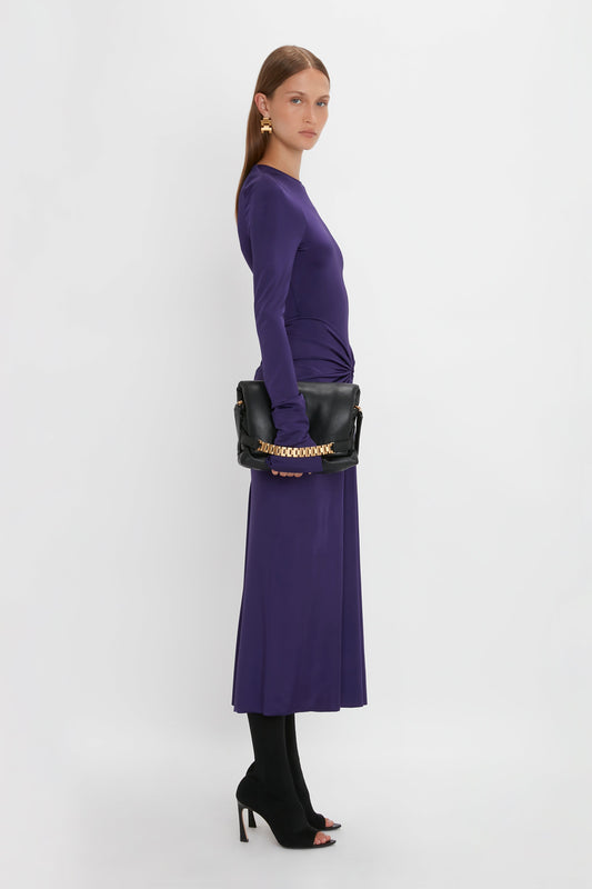 A woman stands sideways wearing the Long Sleeve Gathered Midi Dress In Ultraviolet by Victoria Beckham with black tights and open-toe heels, holding a black handbag adorned with a gold chain. The circular gathered detail of the dress complements its body-sculpting stretch fabric against the plain white background.