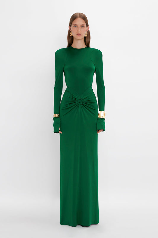 A person stands against a plain background wearing the Victoria Beckham Circle Detail Open Back Gown In Emerald with gold cuff bracelets on both wrists, showcasing an elegant structured silhouette.