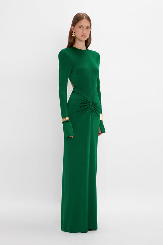 A person with long hair is wearing the Victoria Beckham Circle Detail Open Back Gown In Emerald with gold accessories. The structured silhouette of the gown fits elegantly as they stand against a plain white background.