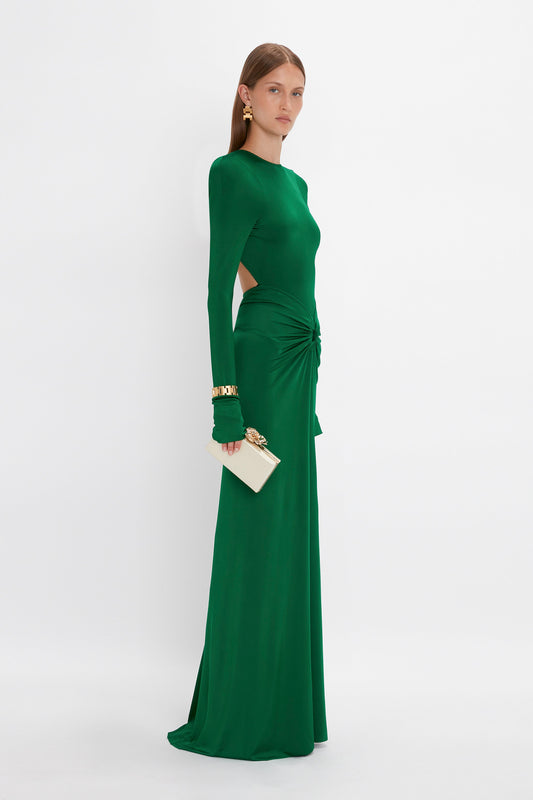 A woman stands in profile wearing the Circle Detail Open Back Gown In Emerald by Victoria Beckham. The structured silhouette is complemented by a small white clutch and gold jewelry.