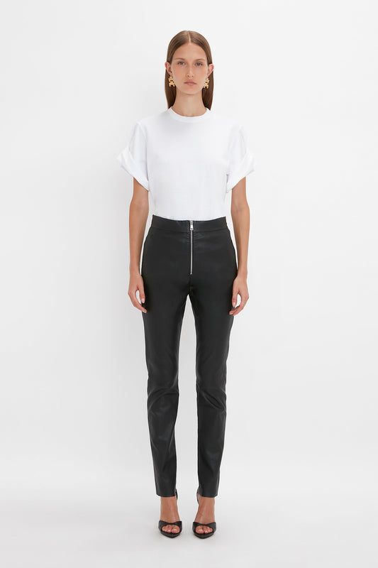 A woman is standing against a plain white background, wearing a white short-sleeved top with rolled cuffs, Victoria Beckham Slim Leather Trouser in Black as an alternative to denim, and black open-toe heels.