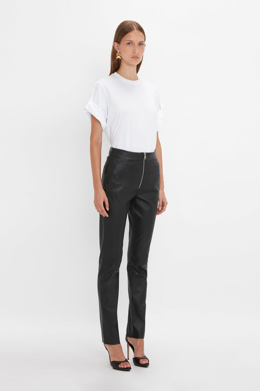 Person standing against a white background, wearing a white rolled-sleeve t-shirt, high-waisted **Slim Leather Trouser in Black** from **Victoria Beckham**, black high-heeled sandals, and gold hoop earrings—a chic alternative to denim.