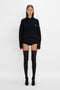 A woman in a black turtleneck sweater and Victoria Beckham Exclusive Over The Knee Socks in Black stands facing the camera on a white background.