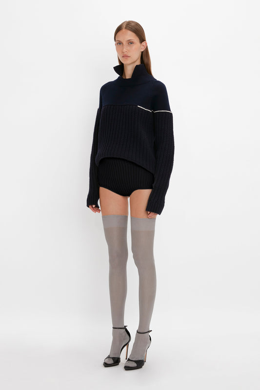 A woman in a studio poses wearing a black oversized sweater, Victoria Beckham Exclusive Over The Knee Socks In Grey, and black high heels.