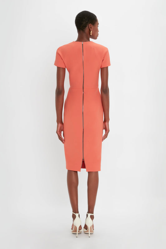 A woman in a coral pink Victoria Beckham Fitted T-Shirt Dress In Papaya with a back zip standing against a white background, viewed from the back.