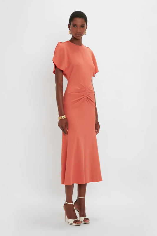 A woman in a Victoria Beckham Gathered Waist Midi Dress In Papaya with a twisted detail at the waist and short sleeves, paired with white pointy-toe stiletto heels, standing against a plain white background.