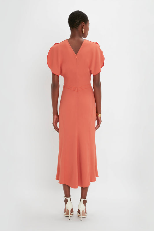 A woman stands facing away, wearing an elegant Victoria Beckham Gathered Waist Midi Dress In Papaya with puff sleeves and white pointy toe stilettos against a plain white background.