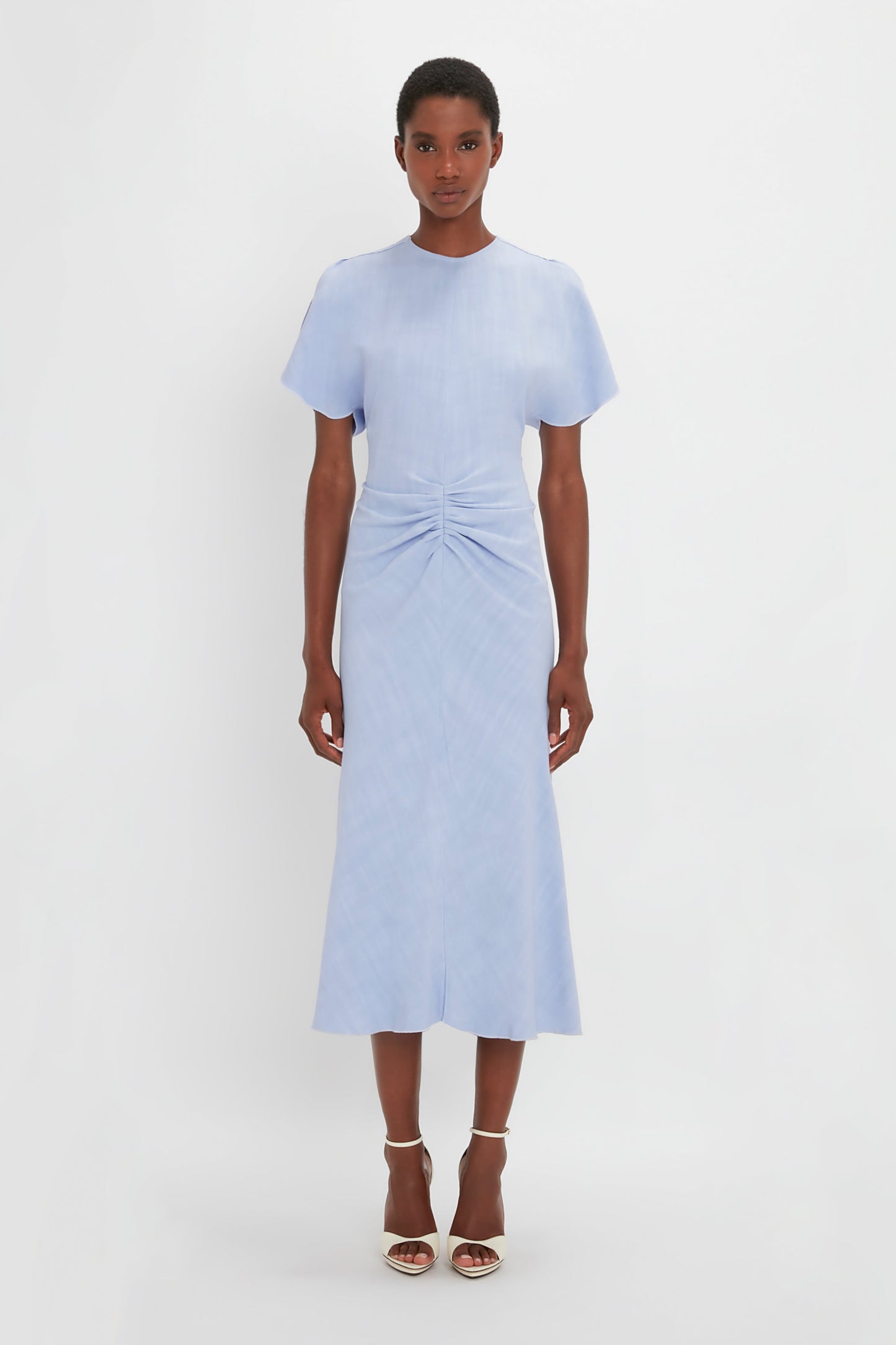 A black woman stands against a white background, wearing a light blue, short-sleeved Gathered Waist Midi Dress In Frost by Victoria Beckham and white pointy toe stilettos.