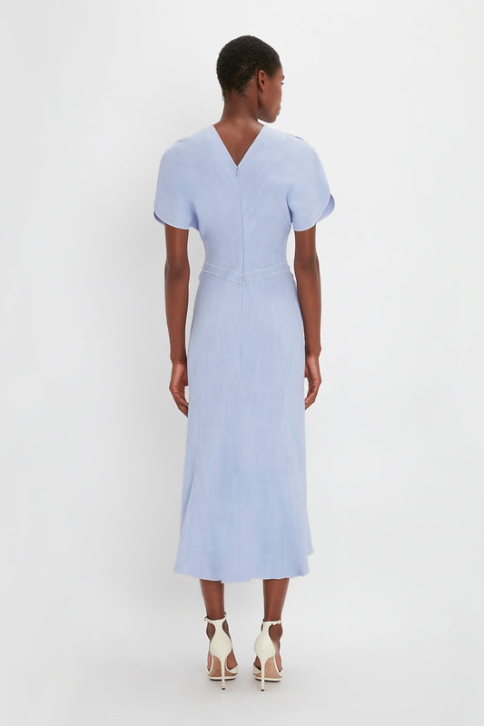 A woman in a Victoria Beckham light blue Gathered Waist Midi Dress In Frost and white pointy toe stilettos, viewed from the back against a plain white background.