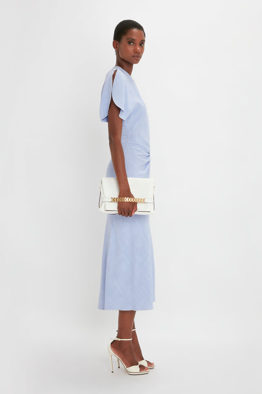 A woman in a Victoria Beckham light blue Gathered Waist Midi Dress In Frost and white pointy toe stiletto heels carries a white handbag, standing sideways and looking at the camera against a gray background.