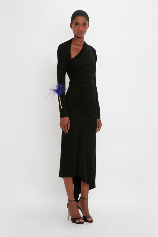 A woman in a Victoria Beckham Slash-Neck Ruched Midi Dress In Black, posing against a plain background, accessorized with a feathered wristband and black heels.