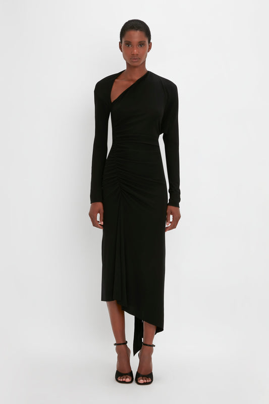 A woman in a Victoria Beckham elegant black gown with an asymmetric cut-out and ruched details, paired with black strappy heels, stands against a white background.