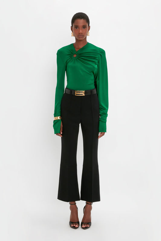 A person is standing against a plain white background wearing a green blouse with a circular detail, **Cropped Kick Trouser In Black by Victoria Beckham**, black heeled sandals, gold earrings, and a gold bracelet.