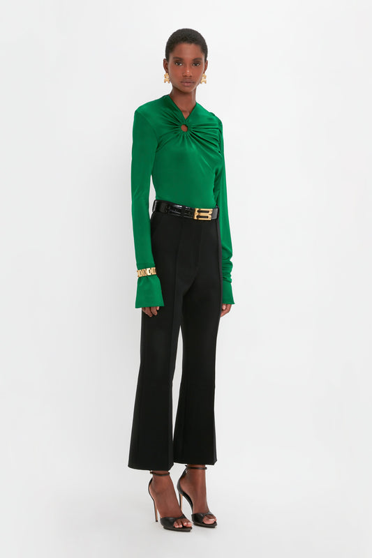 Person wearing a green long-sleeve top with a front ring detail, Cropped Kick Trouser In Black from Victoria Beckham, black belt, black high-heeled sandals, and gold bracelets, standing against a white background.