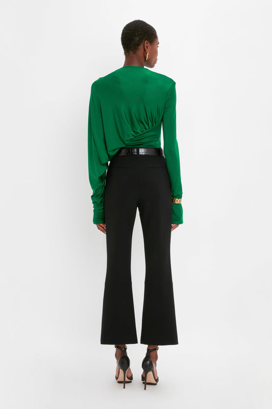 A person stands facing away, wearing a green long-sleeve top and Victoria Beckham's Cropped Kick Trouser In Black. They are also wearing black high heels, and their hair is closely cropped.