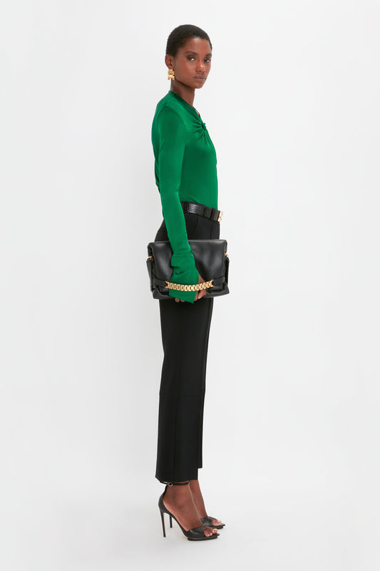 A person wearing a green blouse, Cropped Kick Trouser In Black by Victoria Beckham, and black heels holds a large black handbag with a gold chain strap while standing against a white background.