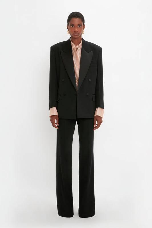 A person stands against a plain background, wearing a Victoria Beckham Satin Lapel Tuxedo Jacket in Black with matching trousers. The individual also has a light-colored collared shirt underneath the suit jacket.