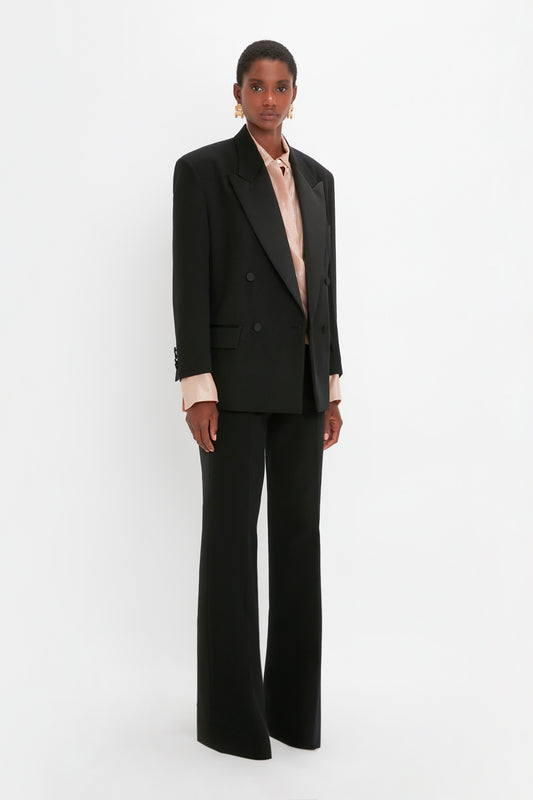 A person stands against a plain background, wearing a Victoria Beckham Satin Lapel Tuxedo Jacket in Black, a light-colored blouse, and black wide-leg pants.