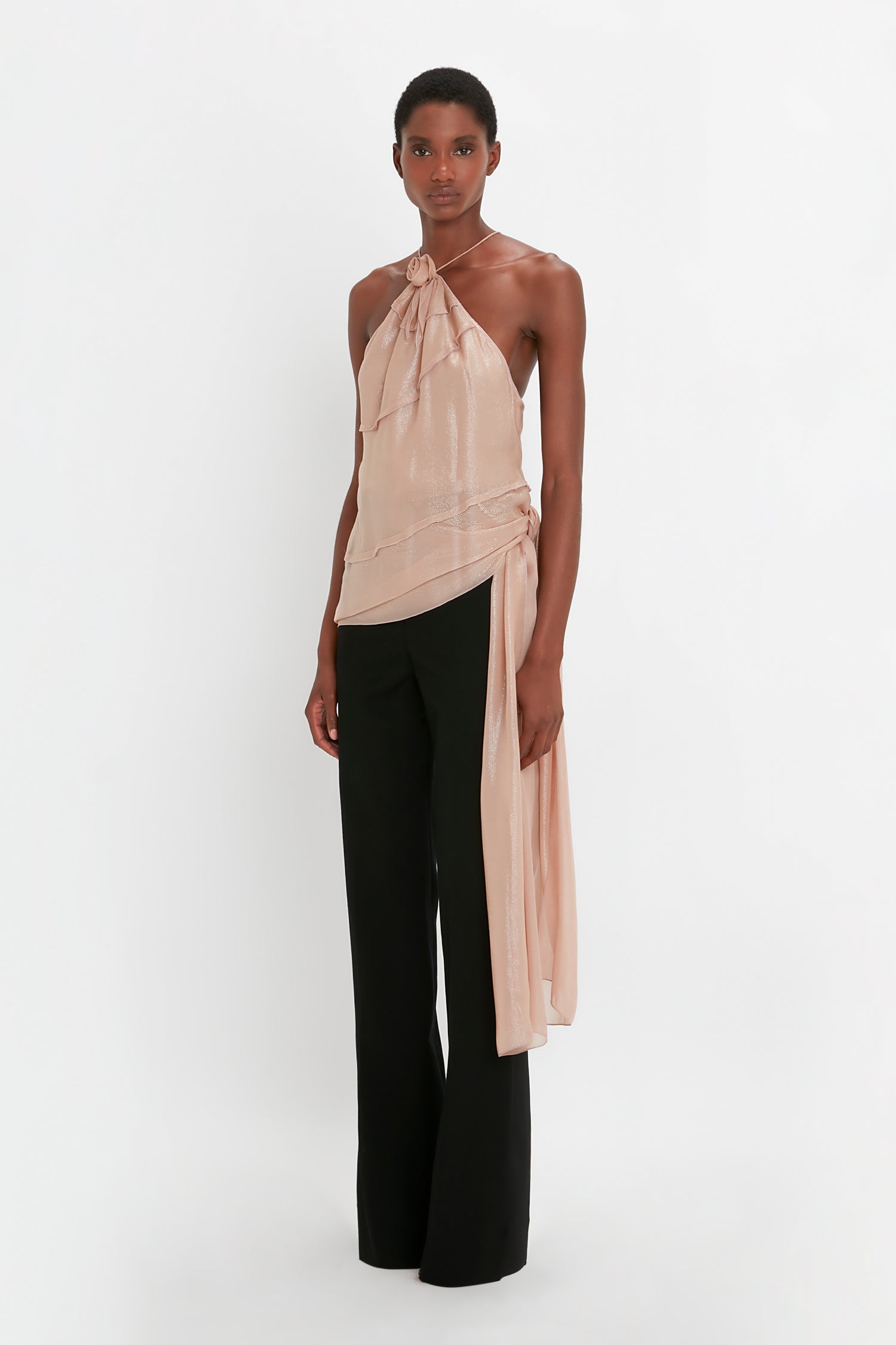 A person in a beige Flower Detail Cami Top In Rosewater by Victoria Beckham, paired with black Satin Panel Straight Leg Trousers, stands against a plain white background.