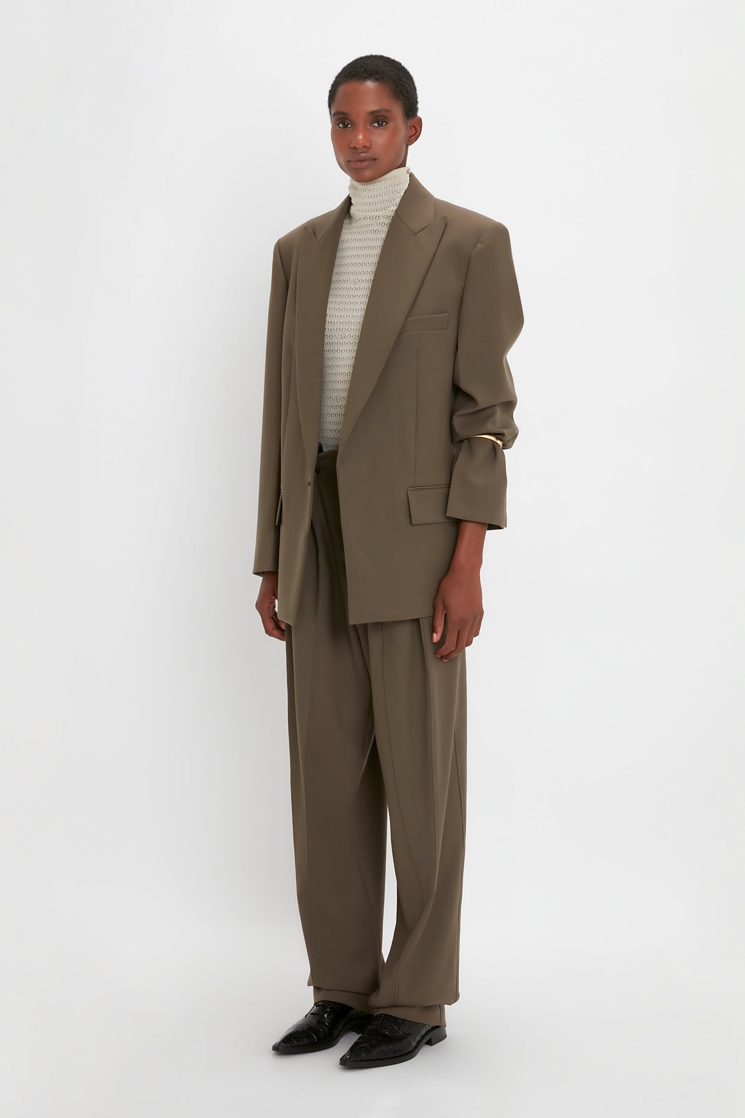 A person stands against a plain background wearing an oversized brown suit with a Peak Lapel Jacket in Oregano by Victoria Beckham, paired with a light turtleneck and black shoes, exuding a contemporary aesthetic.