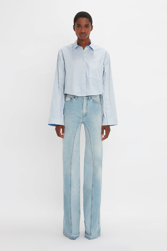 Person standing against a plain background, wearing a Victoria Beckham Button Detail Cropped Shirt In Chamomile Blue Stripe and light blue high-waisted jeans, showcasing a sophisticated yet casual feminine silhouette.