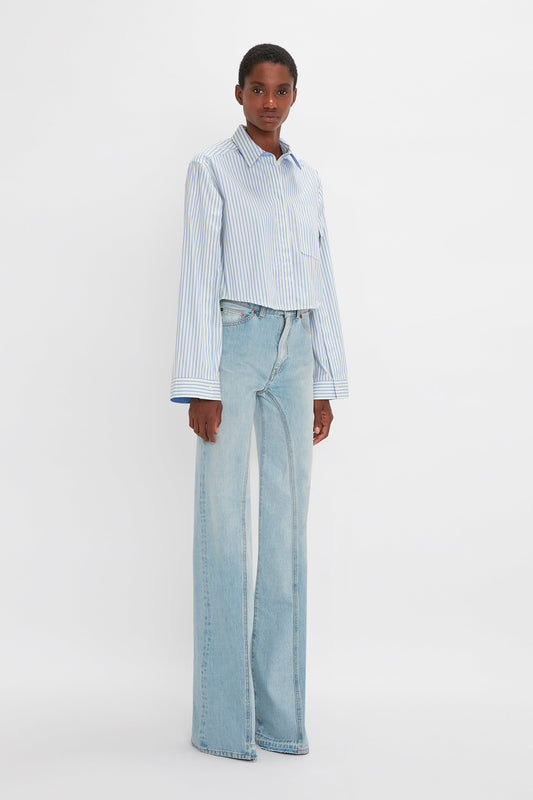 A person stands against a plain white background wearing a Victoria Beckham Button Detail Cropped Shirt In Chamomile Blue Stripe and light blue wide-leg jeans, showcasing a feminine silhouette. They look directly at the camera.