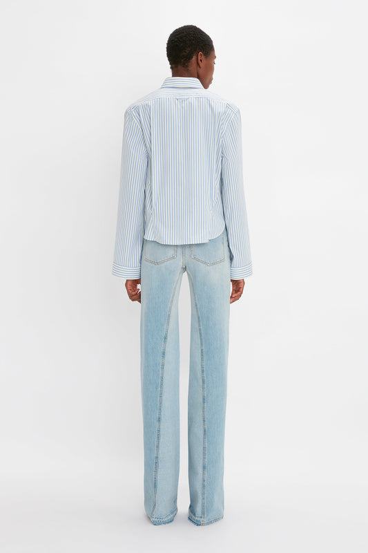 A person with short hair is standing and facing away from the camera, wearing a blue and white striped Button Detail Cropped Shirt In Chamomile Blue Stripe by Victoria Beckham, paired with light blue jeans. The background is plain white.