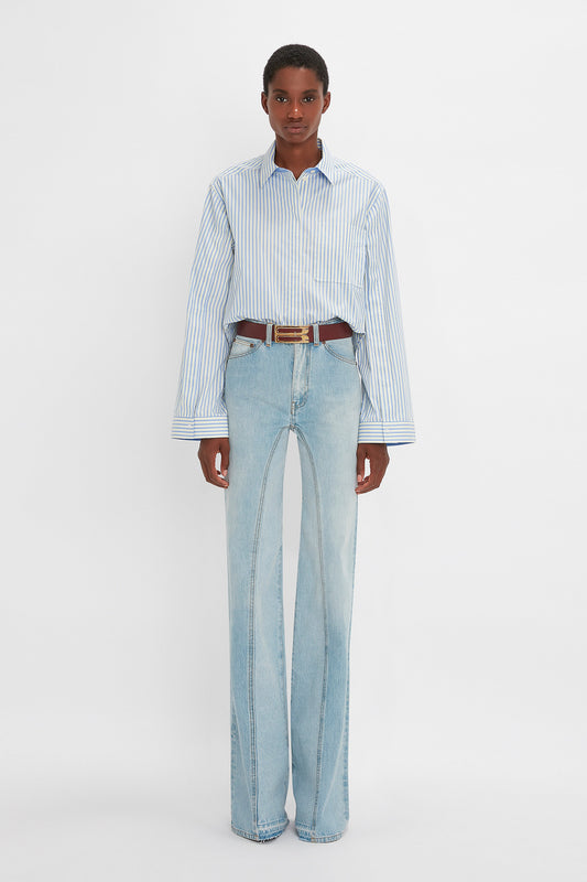 A man standing in a studio wearing a striped blue and white button detail cropped shirt, wide-leg Victoria Beckham Bianca jeans, and a brown belt, with a neutral expression and hands at his sides.