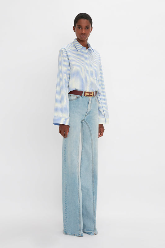 A woman stands in a studio wearing a striped button detail cropped shirt, Victoria Beckham Light Blue Denim flared jeans with a contrasting belt, and facing the camera with a subtle expression.