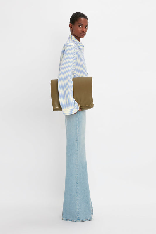 A person wearing a Victoria Beckham Button Detail Cropped Shirt In Chamomile Blue Stripe, light blue flared jeans, and holding a large olive-green clutch stands against a white background, showcasing a feminine silhouette.