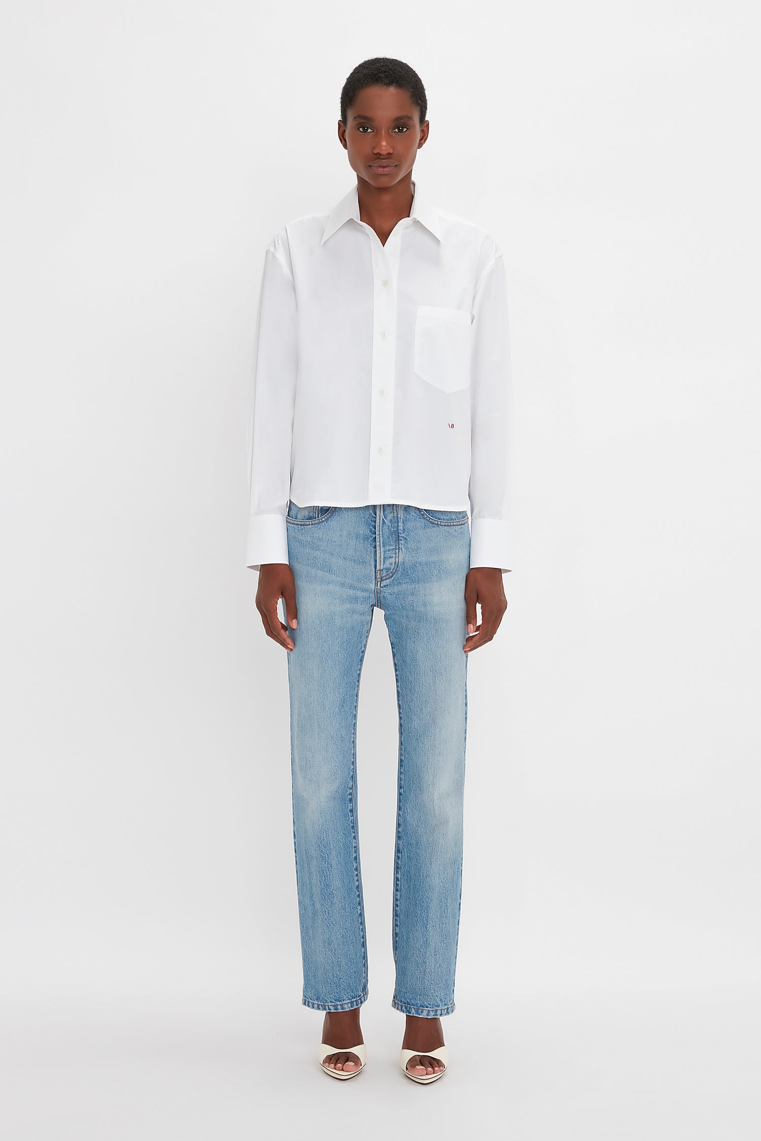 An individual wearing a Victoria Beckham Cropped Long Sleeve Shirt In White, blue jeans, and open-toe sandals stands against a plain white background.