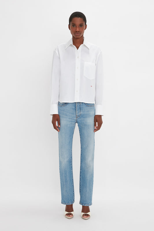 An individual wearing a Victoria Beckham Cropped Long Sleeve Shirt In White, blue jeans, and open-toe sandals stands against a plain white background.