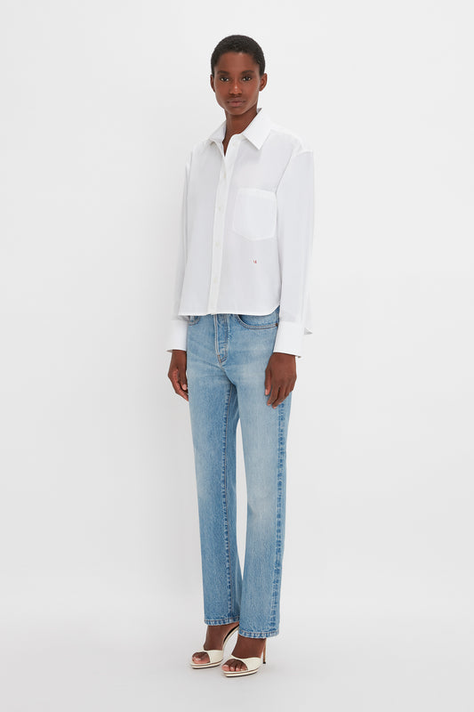 Person standing against a white background, wearing the Cropped Long Sleeve Shirt In White by Victoria Beckham, light blue jeans, and white heeled sandals. They have short hair and a neutral expression.