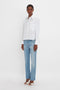 Person standing against a white background, wearing the Cropped Long Sleeve Shirt In White by Victoria Beckham, light blue jeans, and white heeled sandals. They have short hair and a neutral expression.