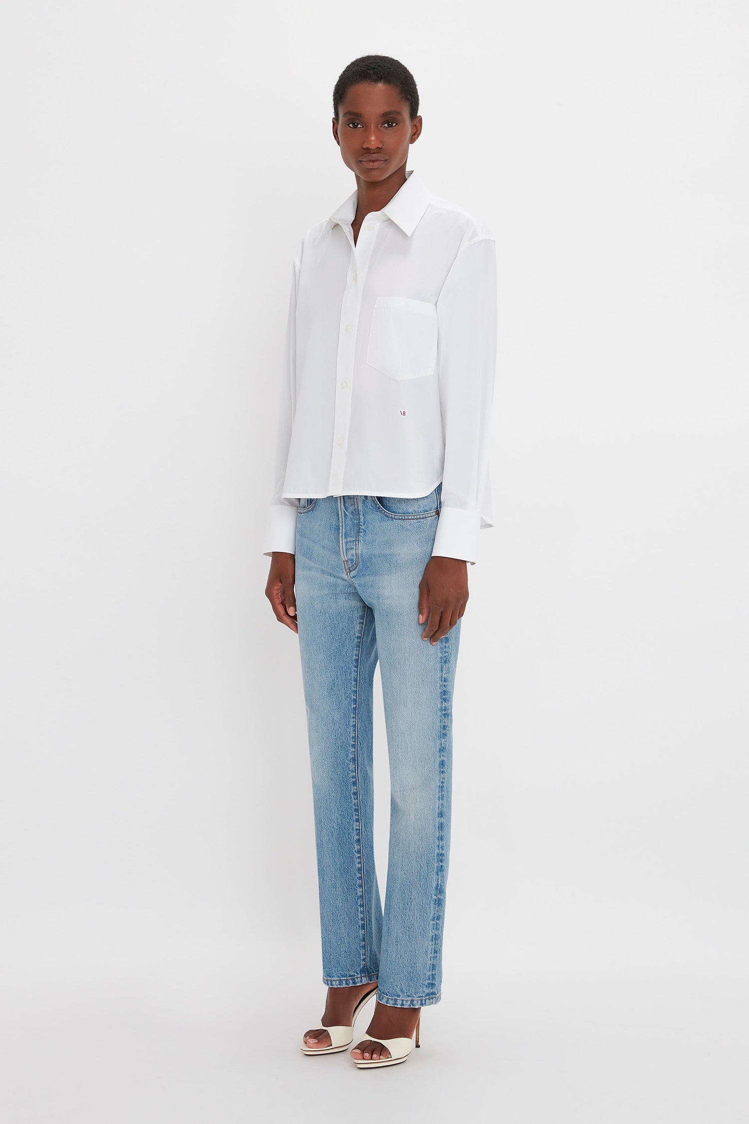 A woman wearing a classic Victoria Beckham cropped long sleeve shirt in white and blue jeans standing against a plain white background.
