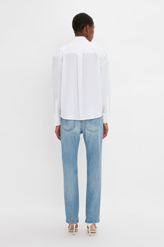 Person standing with their back to the camera, wearing a Cropped Long Sleeve Shirt In White by Victoria Beckham, blue jeans, and white heeled sandals, against a plain white background. The relaxed tailoring evokes an effortless elegance reminiscent of Victoria Beckham's signature style.