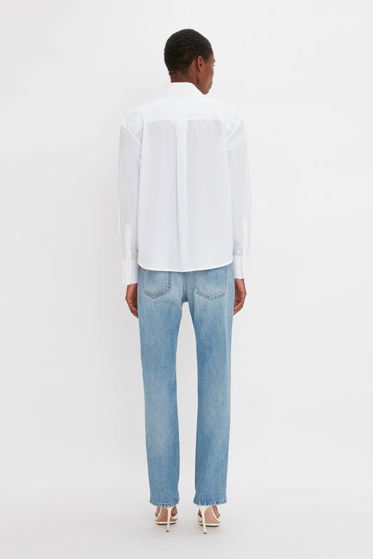 A rear view of a woman standing, wearing a Victoria Beckham cropped long sleeve shirt in white with pleat detail and blue jeans, styled with white high-heeled shoes.