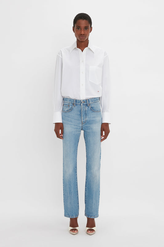 A person stands against a white background wearing a white button-up shirt, Victoria Mid-Rise Jean In Light Blue by Victoria Beckham, and shoes with small heels.
