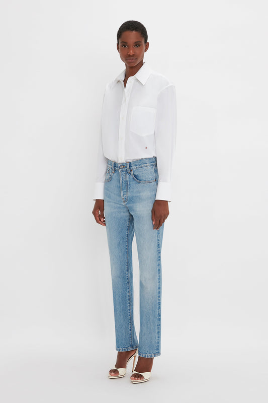 A person stands against a plain white background, wearing a white button-up shirt, Victoria Beckham Victoria Mid-Rise Jean In Light Blue, and white high-heeled sandals.