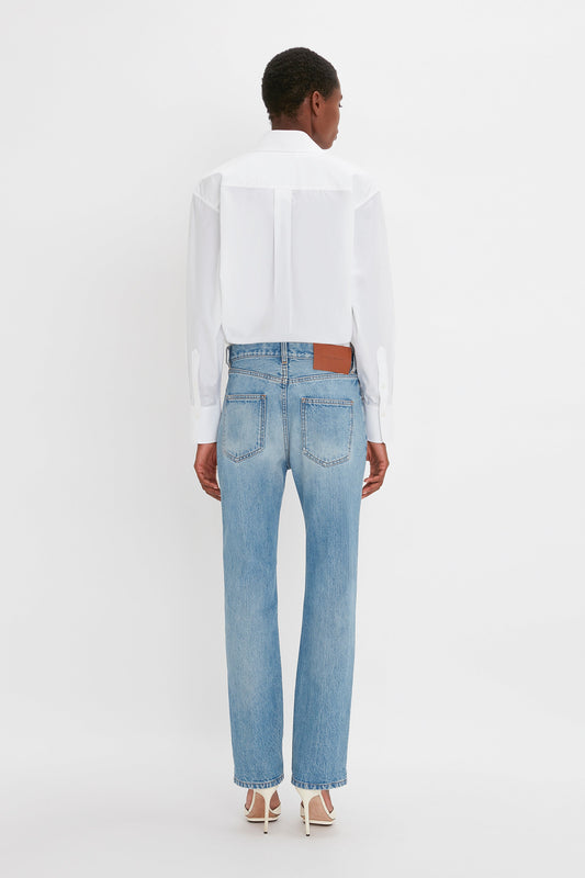 A person stands with their back to the camera, wearing a white shirt, Victoria Mid-Rise Jean In Light Blue by Victoria Beckham, and white heeled shoes against a plain white background.