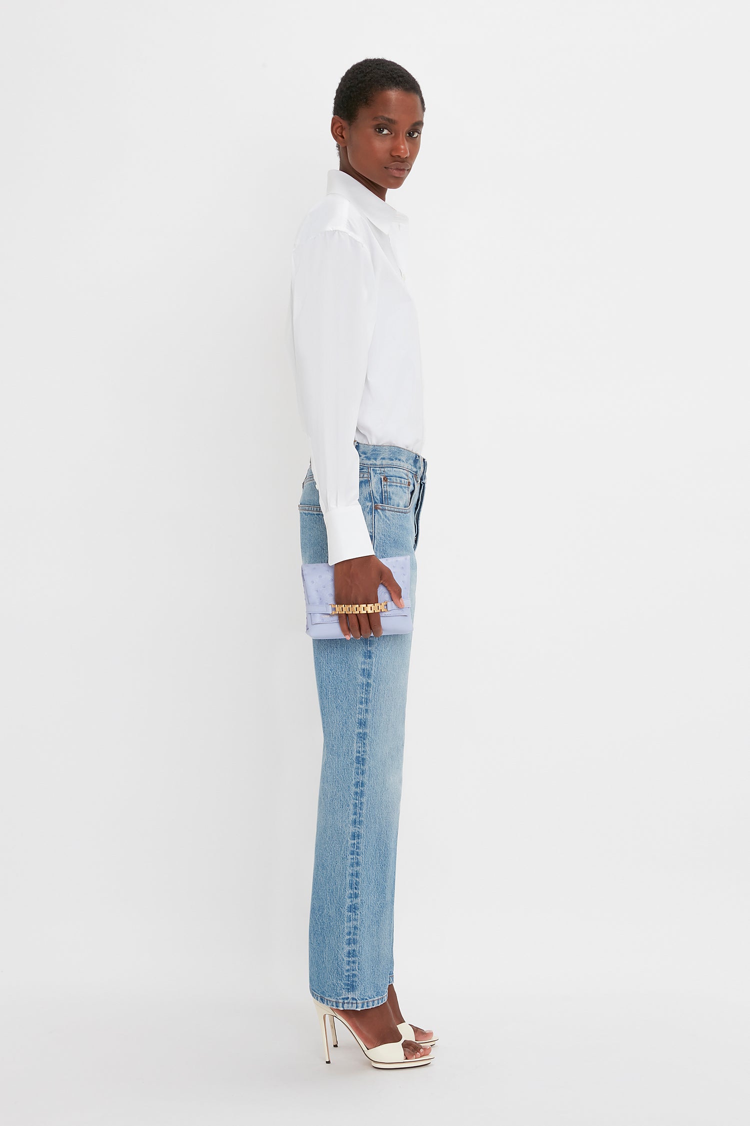 Person in a Victoria Beckham Cropped Long Sleeve Shirt In White and blue jeans holds a purple clutch, standing sideways. They wear white high-heeled shoes. Background is plain white.
