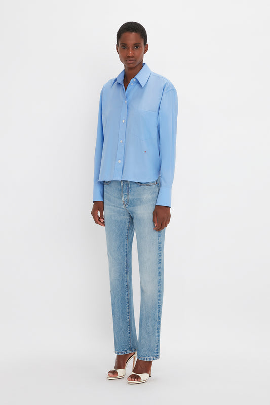 A person stands against a plain white background, wearing a relaxed fit light blue *Cropped Long Sleeve Shirt In Oxford Blue* by *Victoria Beckham*, light blue jeans, and white high-heeled shoes.