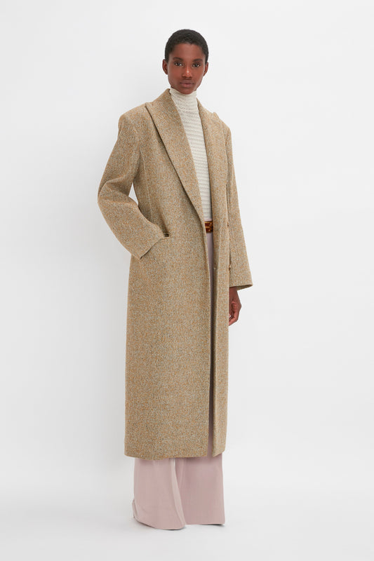 A person stands against a white background, showcasing an Exclusive Waisted Tailored Coat In Flax by Victoria Beckham: a long beige double-breasted coat over a white turtleneck and light pink trousers, exemplifying precision tailoring.