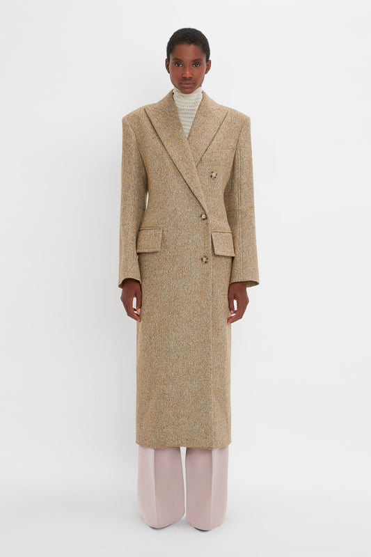 Person wearing a Victoria Beckham **Exclusive Waisted Tailored Coat In Flax**—a long beige double-breasted coat with structured shoulders—paired with a light gray turtleneck and wide-leg white trousers, standing against a plain white background.